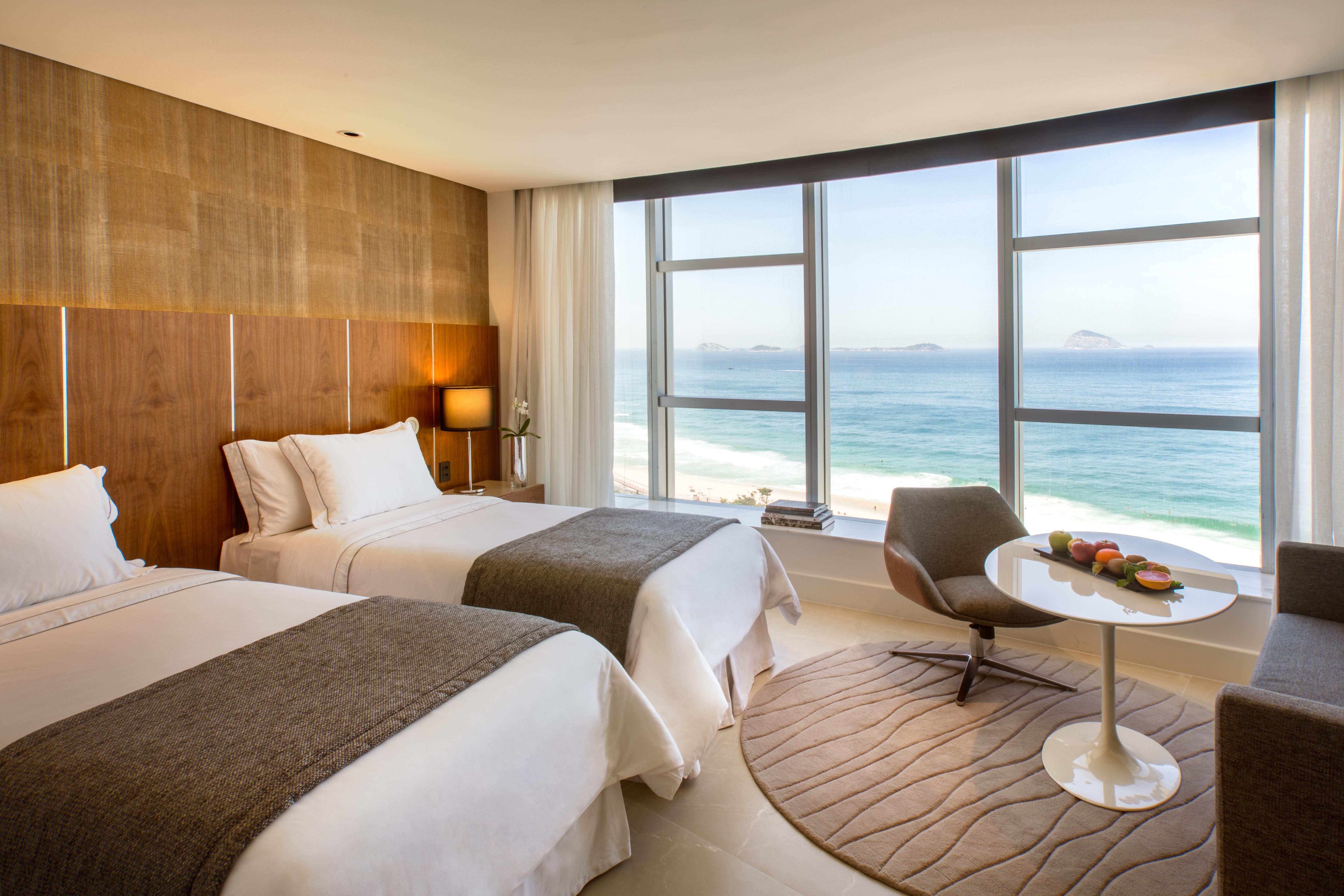 Hotel Nacional Rio De Janeiro - Oficial Esterno foto The photo shows a modern hotel room with two beds adorned in white linens and gray throws. Large windows allow natural light to flood the space, offering a stunning view of the ocean. In the corner, there's a comfortable chair and a small round table